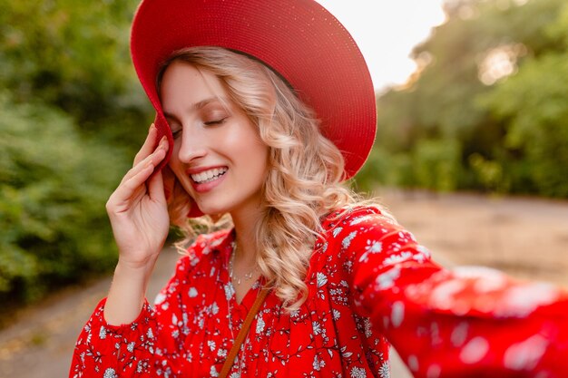 Mujer sonriente rubia elegante atractiva en traje de la manera del verano del sombrero rojo de la paja y de la blusa que toma la foto del selfie