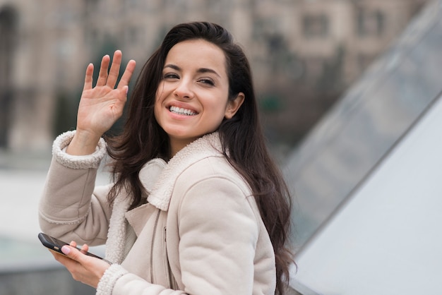 Foto gratuita mujer sonriente saludando mientras sostiene el teléfono inteligente