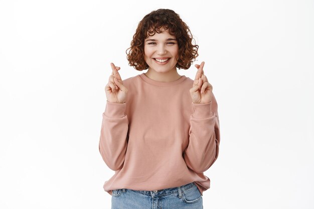 Una mujer sonriente y segura de sí misma cruza los dedos para tener buena suerte haciendo que el deseo se vea determinado y asertivo cuando reza de pie contra el fondo blanco Copiar espacio