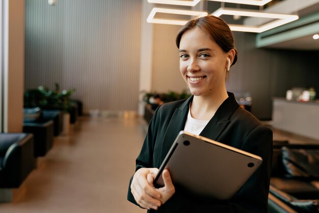 Una mujer sonriente y segura de sí misma con el pelo recogido, con una chaqueta oscura y una camiseta blanca, sostiene una laptop y sonríe mientras trabaja en la oficina