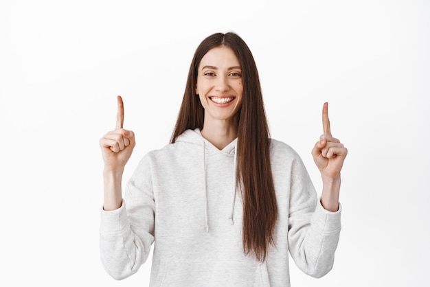 Mujer sonriente segura de sí misma recomendando hacer clic en el logotipo en el banner del sitio web apuntando con el dedo hacia arriba y mirando a la cámara que muestra la mejor oferta en la tienda de pie sobre fondo blanco