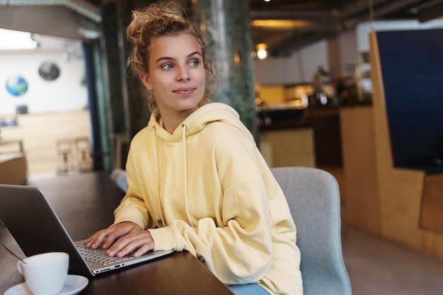 Mujer sonriente sentada en un café con una computadora portátil mirando por la ventana complacida Mujer independiente creativa inspirándose trabajando al aire libre en una cafetería Chica estudiando en línea en un café con una computadora portátil