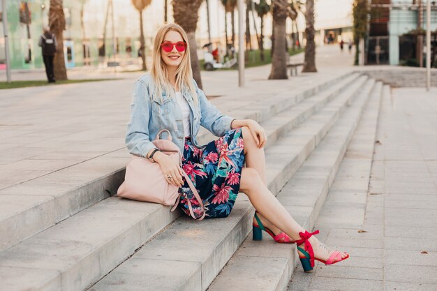 Mujer sonriente sentada en las escaleras en las calles de la ciudad con elegante falda estampada y chaqueta vaquera de gran tamaño con mochila de cuero con gafas de sol rosa