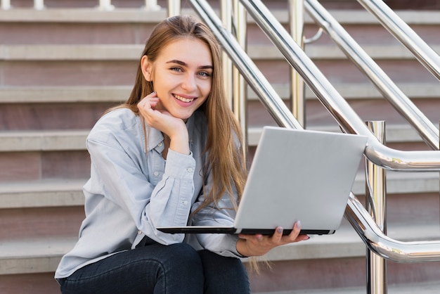 Mujer sonriente sentada en las escaleras y sosteniendo una computadora portátil