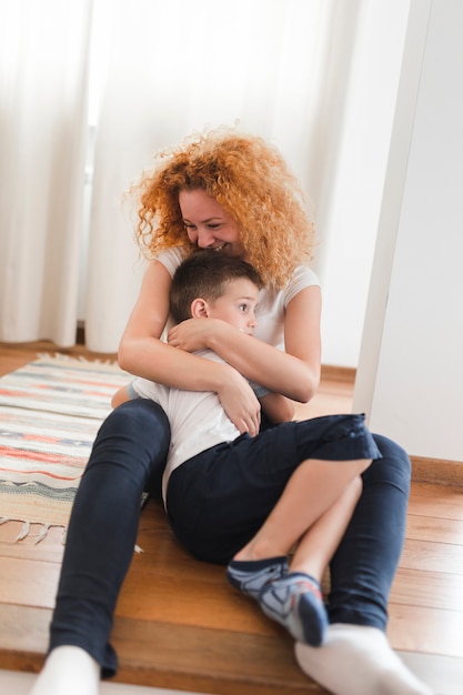 Foto gratuita mujer sonriente sentada en el piso de madera dura abrazando a su hijo