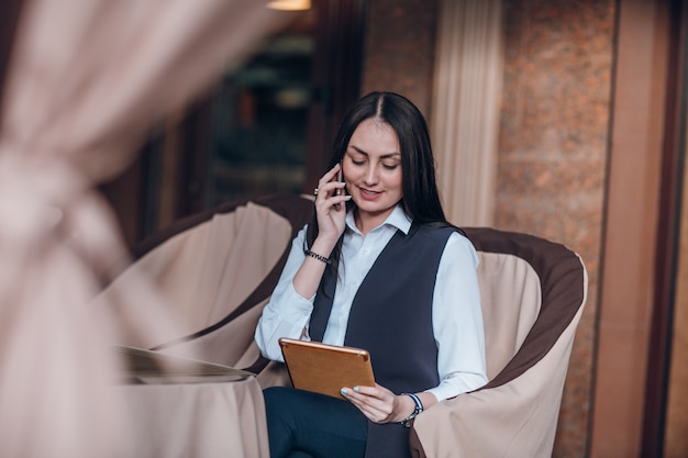 Mujer sonriente sentada en un restaurante elegante con una tablet y hablando por teléfono