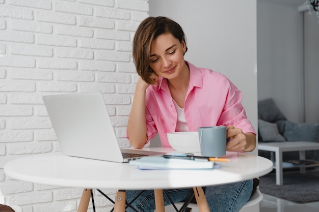 Mujer sonriente sincera en camisa rosa desayunando en casa en la mesa trabajando en línea en la computadora portátil desde casa, comiendo cereales