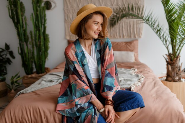 Mujer sonriente en sombrero de paja que se enfría en casa en el acogedor interior de boho