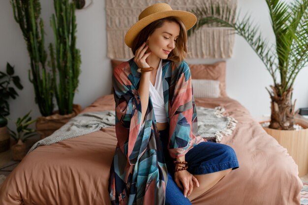 Mujer sonriente en sombrero de paja que se enfría en casa en el acogedor interior de boho