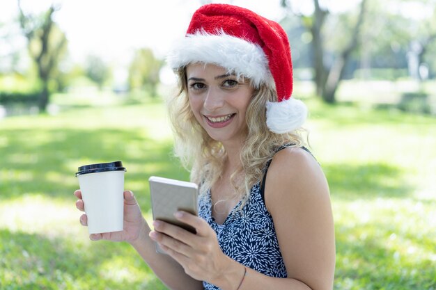 Mujer sonriente con sombrero de Santa, sosteniendo café y teléfono inteligente