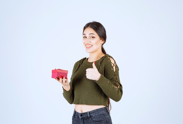 Mujer sonriente sosteniendo caja de regalo y dando pulgar hacia arriba sobre fondo blanco.