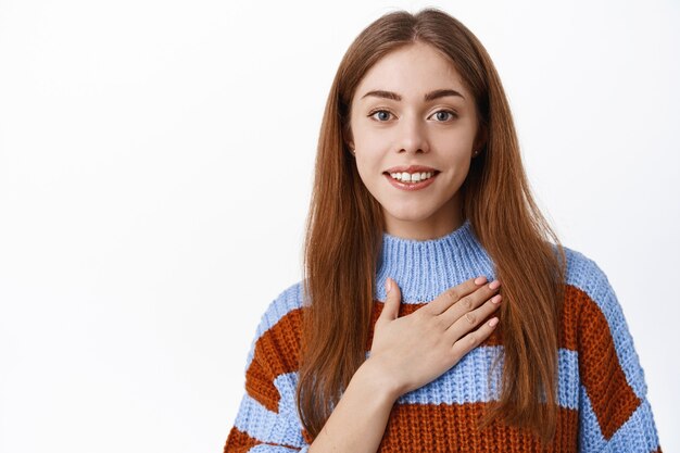 Mujer sonriente sosteniendo la mano en el corazón y diciendo gracias, agradecida por algo, mira con gratitud y rostro sincero, pared blanca