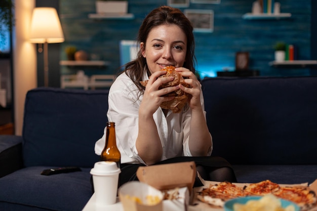 Mujer sonriente sosteniendo sabrosa hamburguesa en las manos mirando comedia de situación de comedia de televisión de pie en el sofá frente a la mesa con menú de comida rápida para llevar. Persona feliz viendo el programa de televisión cenando para llevar.