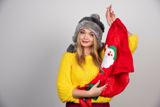 Mujer sonriente en suéter amarillo y sombrero sosteniendo una bolsa de Navidad roja.