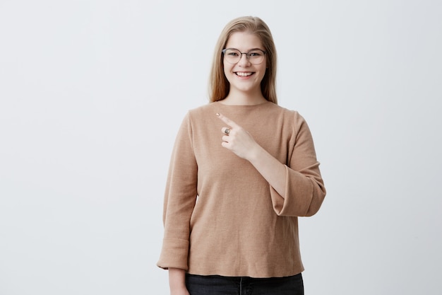 Mujer sonriente en suéter y gafas con estilo con el pelo rubio liso apuntando a la pared blanca en blanco, demostrando algo. Hembra alegre que indica con el dedo índice en fondo gris.