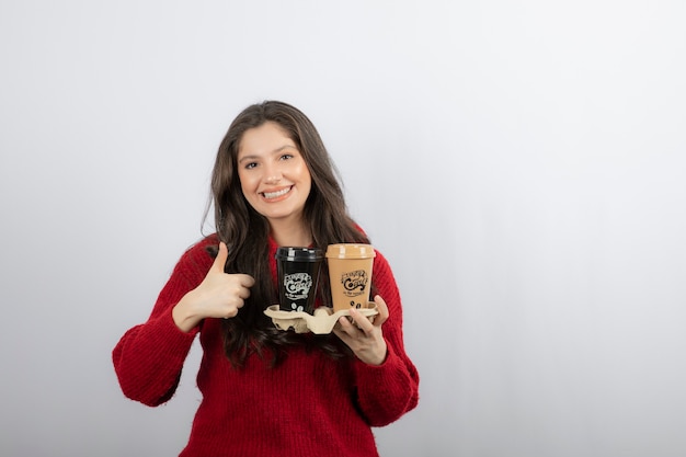 Mujer sonriente con tazas de café en el soporte de cartón mostrando un pulgar hacia arriba.