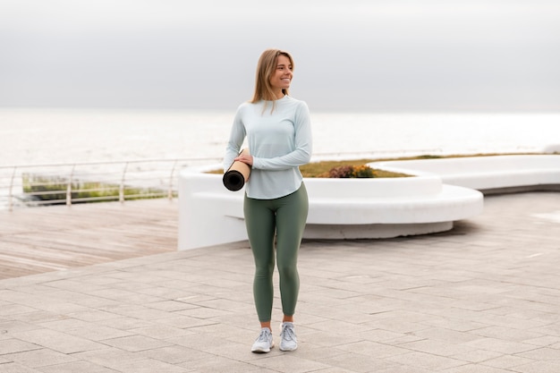 Foto gratuita mujer sonriente de tiro completo sosteniendo estera de yoga