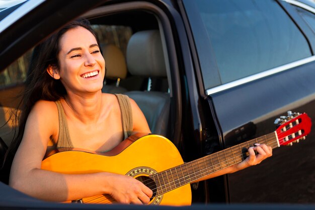 Mujer sonriente tocando la guitarra fuera de su coche mientras está al aire libre