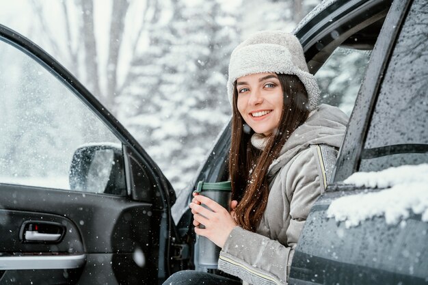 Mujer sonriente tomar una bebida caliente y disfrutar de la nieve durante un viaje por carretera