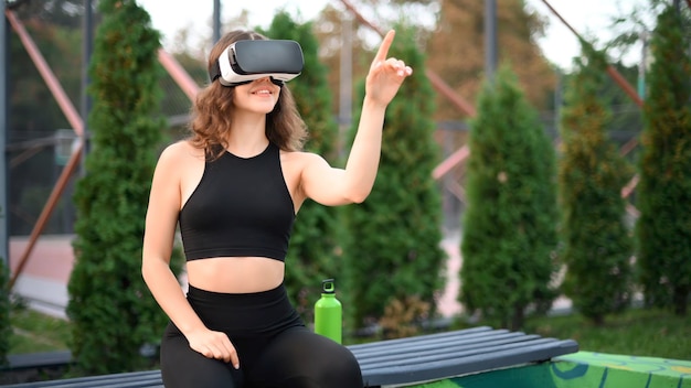 Foto gratuita mujer sonriente usando auriculares vr en un chándal mientras está sentada en un banco en un campo deportivo en un parque