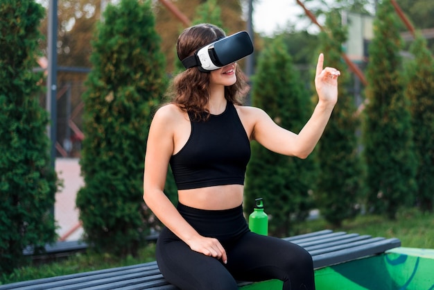 Foto gratuita mujer sonriente usando auriculares vr en un chándal mientras está sentada en un banco en un campo deportivo en un parque