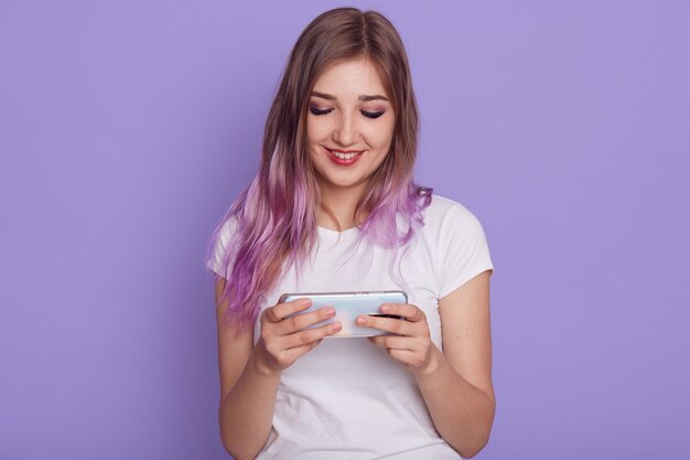 La mujer sonriente viste la camiseta blanca con el teléfono inteligente en las manos, juego de chapado, mirando la pantalla con una linda sonrisa, usando el teléfono celular, posando aislado sobre la pared púrpura.