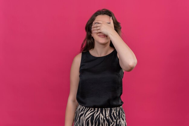 Mujer sonriente vistiendo camiseta negra cubrió su rostro con la mano en la pared rosa