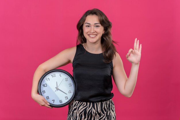 Mujer sonriente vistiendo camiseta negra con reloj de pared y muestra gesto okey en pared rosa