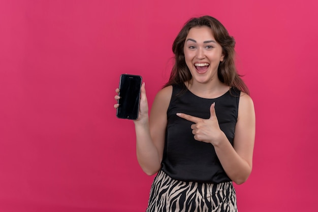 Mujer sonriente vistiendo camiseta negra sosteniendo un teléfono y apuntar el teléfono en la pared rosa
