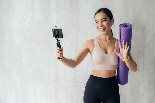 Mujer sonriente vlogueando con su teléfono mientras sostiene una colchoneta de fitness