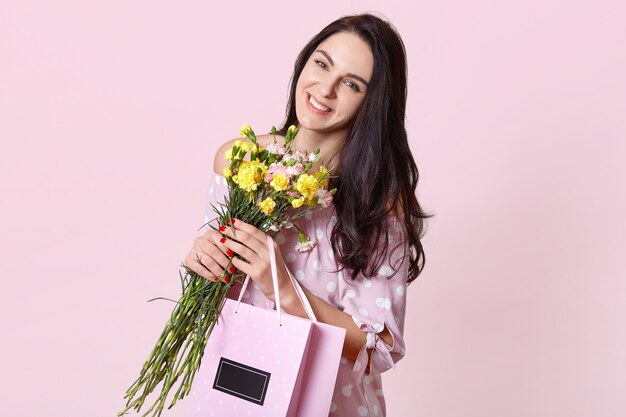 mujer con una sonrisa con dientes, tiene el pelo largo y liso y oscuro, inclina la cabeza, lleva un ramo de flores y una bolsa con el presente, posa en rosa, expresa emociones positivas, tiene un buen día