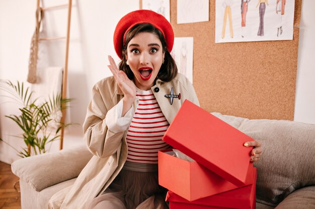 Mujer sorprendida abre caja de regalo y mira a cámara. Chica maravillosa sorprendida en ropa moderna de otoño se regocija con su regalo.