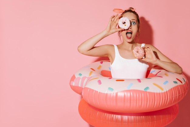 Mujer sorprendida con aretes elegantes en traje de baño fresco y ligero posando con donuts y grandes anillos de natación en un fondo aislado