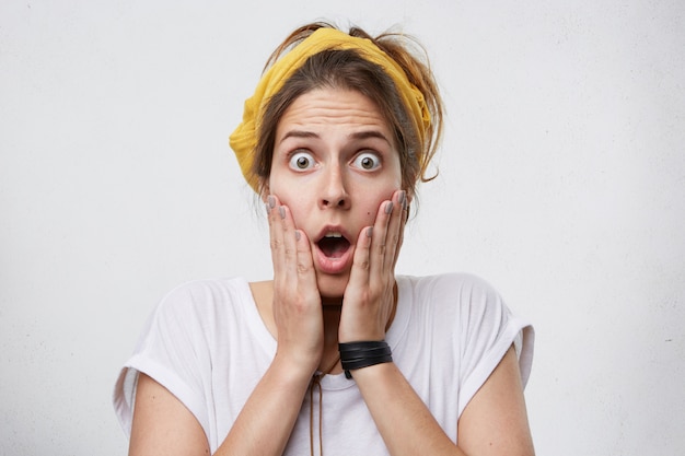 Mujer sorprendida asombrada asustando algo mirando con los ojos bien abiertos y la boca tomados de la mano en las mejillas con camiseta blanca casual. Mujer tiene miedo de las dificultades mirando