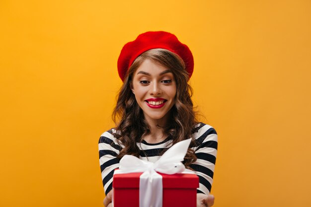 Mujer sorprendida en boina roja y camisa a rayas tiene caja de regalo. Encantadora dama con peinado ondulado con sombrero brillante y blusa moderna posando.