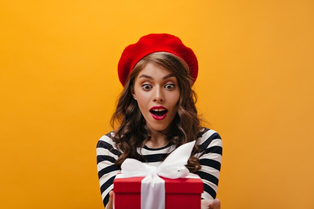 Mujer sorprendida en boina roja recibiendo caja de regalo. Chica joven rizada con lápiz labial brillante en sombrero posando sobre fondo aislado.