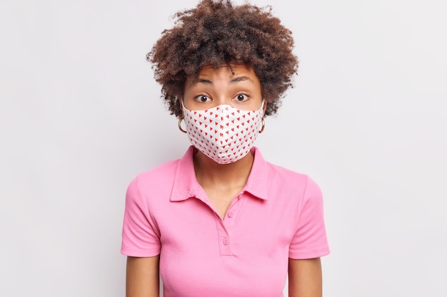 Mujer sorprendida con cabello rizado usa una máscara desechable durante la cuarentena y el brote de coronavirus se preocupa por la salud usa una camiseta rosa informal aislada sobre una pared blanca