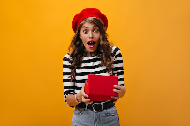 Mujer sorprendida en caja de regalo de apertura de boina roja sobre fondo naranja.Muchacha encantadora con peinado ondulado con sombrero brillante y ropa moderna se regocija.