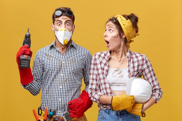 Foto gratuita mujer sorprendida en camisa a cuadros con casco mirando a su marido con la boca abierta que hizo algo mal. hombre joven hermoso asombrado en la máscara protectora que sostiene la perforadora