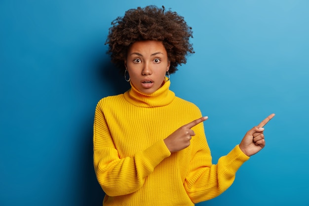 Mujer sorprendida confundida con puntos de corte de pelo afro a un lado, se da cuenta de que sucedió algo sorprendente, tiene expresión ansiosa preocupada aislada sobre fondo azul