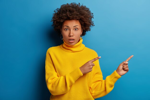 Mujer sorprendida confundida con puntos de corte de pelo afro a un lado, se da cuenta de que sucedió algo sorprendente, tiene expresión ansiosa preocupada aislada sobre fondo azul