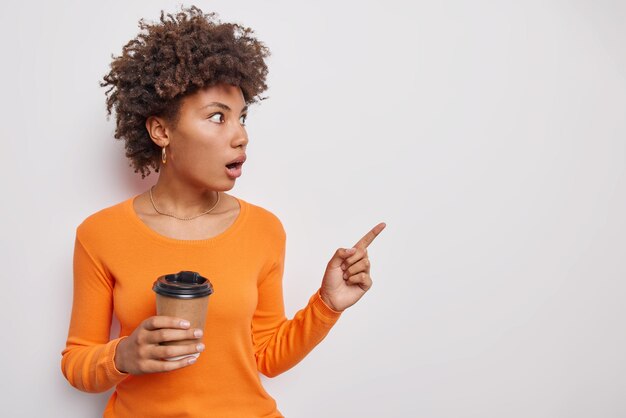 Mujer sorprendida y conmocionada mira emotiva indica con el dedo índice alejado en el espacio en blanco contiene el aliento de asombro bebidas café para llevar vestido con jersey naranja aislado sobre fondo blanco