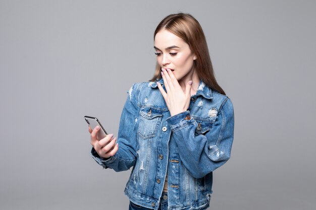 Mujer sorprendida cubriendo la boca, mirando la pantalla del teléfono móvil, sorprendida mujer leyendo un mensaje inesperado, oferta de compra, buenas noticias, sosteniendo el teléfono celular, aislado en la pared gris