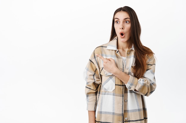 Mujer sorprendida y emocionada dice wow, señalando y mirando a un lado la oferta de promoción súper impresionante, la mejor oferta en la tienda, de pie sobre una pared blanca bajo una gran impresión