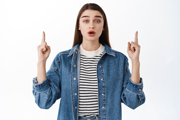 Mujer sorprendida y emocionada hablando sobre un nuevo producto, haciendo un anuncio, mire allí, señalando con el dedo hacia arriba y mirando asombrada, mostrando publicidad, de pie sobre una pared blanca