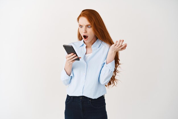Una mujer sorprendida y enojada con el pelo rojo recibe spam en el teléfono, lee un mensaje irritante en el teléfono inteligente y parece indignada