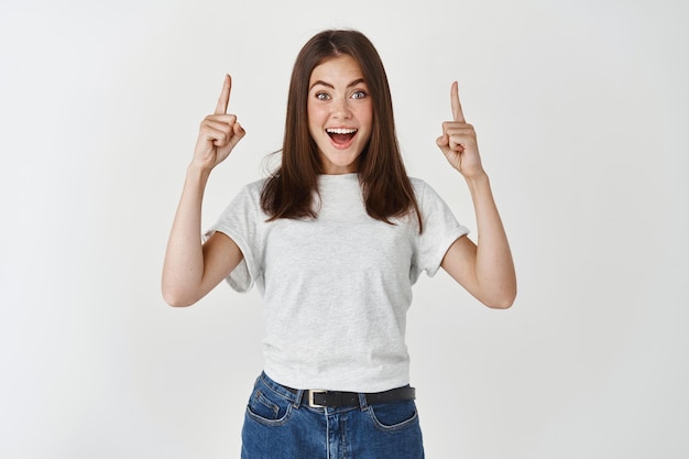 Foto gratuita mujer sorprendida y feliz mostrando publicidad, apuntando con las manos hacia arriba y mirando al frente fascinado, de pie sobre una pared blanca.