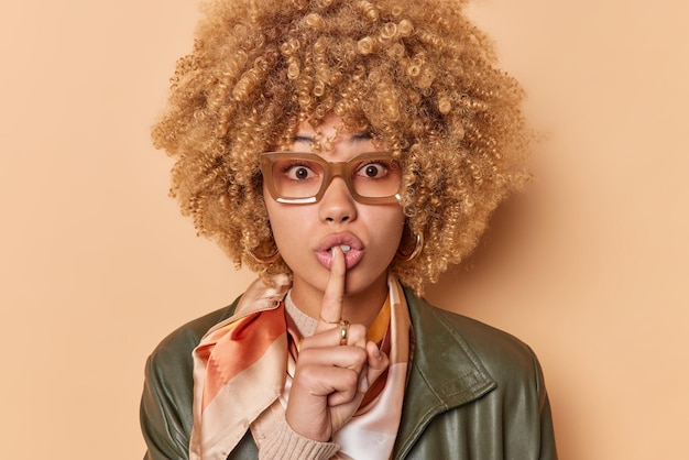 Una mujer sorprendida hace un gesto tabú, un signo de silencio, se calla en el interior, se ve una expresión misteriosa vestida con ropa elegante, usa anteojos aislados sobre un fondo marrón. Shh mantén mi secreto a salvo