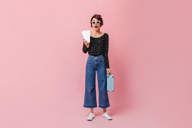 Mujer sorprendida en jeans sosteniendo boletos en pared rosa