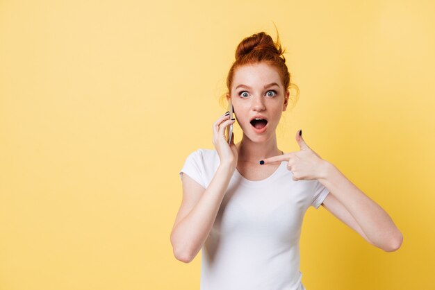 Mujer sorprendida de jengibre en camiseta hablando y señalando en el teléfono inteligente